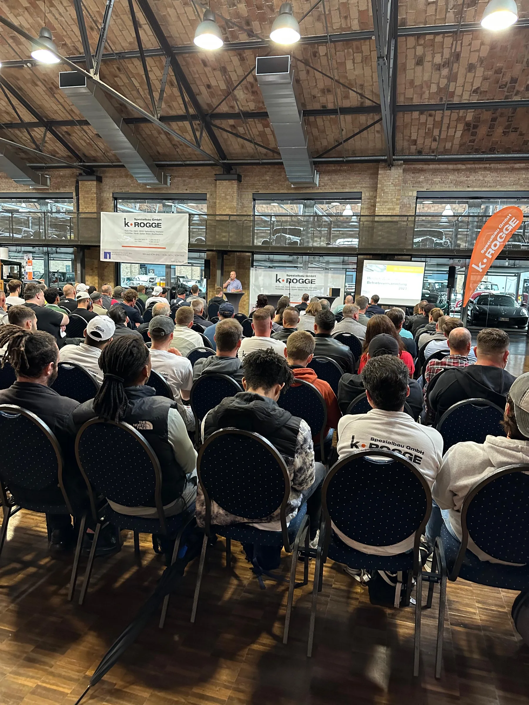 Voll besetzte Stuhlreihen in einer Halle mit Holzdecke, vorne eine Leinwand