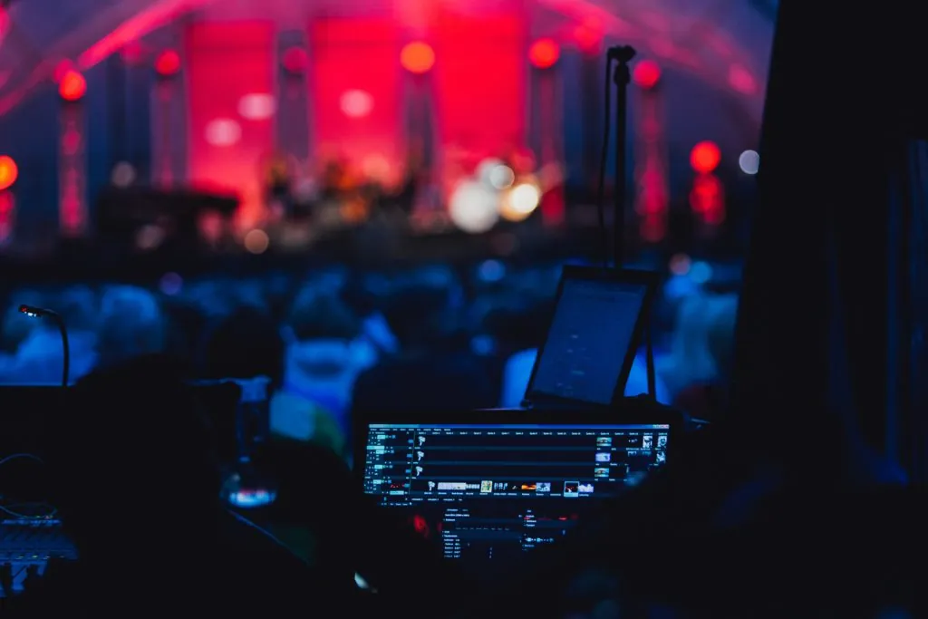 Mischpult mit Bildschirm vor einer rot beleuchteten Bühne bei einem Konzert