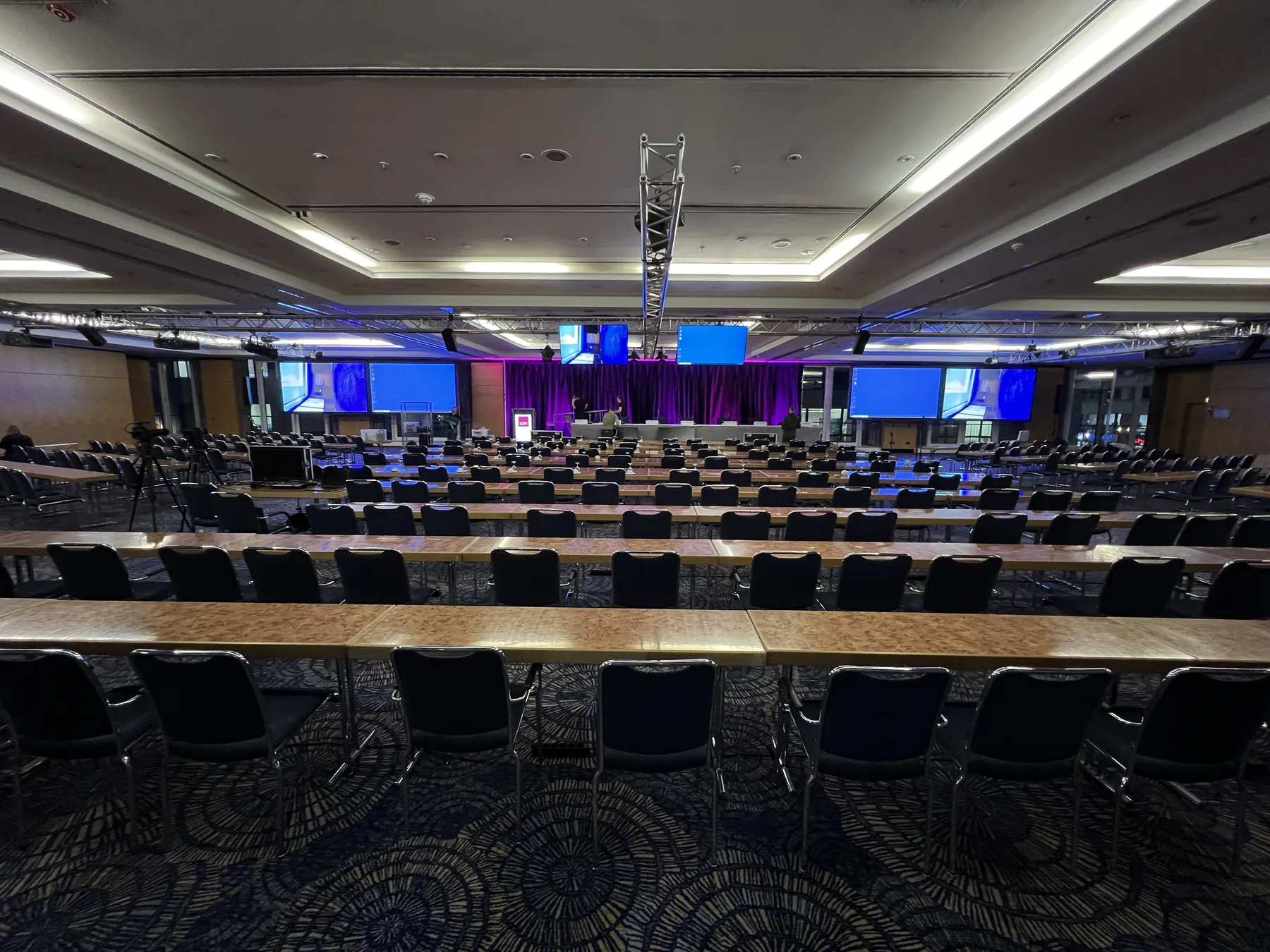 Großer Konferenzsaal mit Tischreihen, Bühne und mehreren Leinwänden, vor Beginn